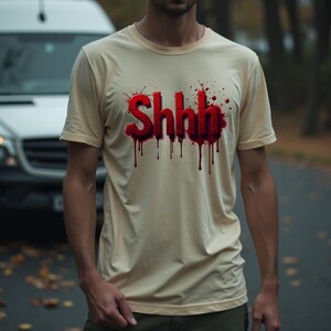 Pu&ograve; includere: T-shirt color crema con la scritta "Shhh" in rosso, con un effetto gocciolante simile a sangue. La maglietta a maniche corte ha uno scollo tondo. La persona indossa la maglietta all'aperto.
