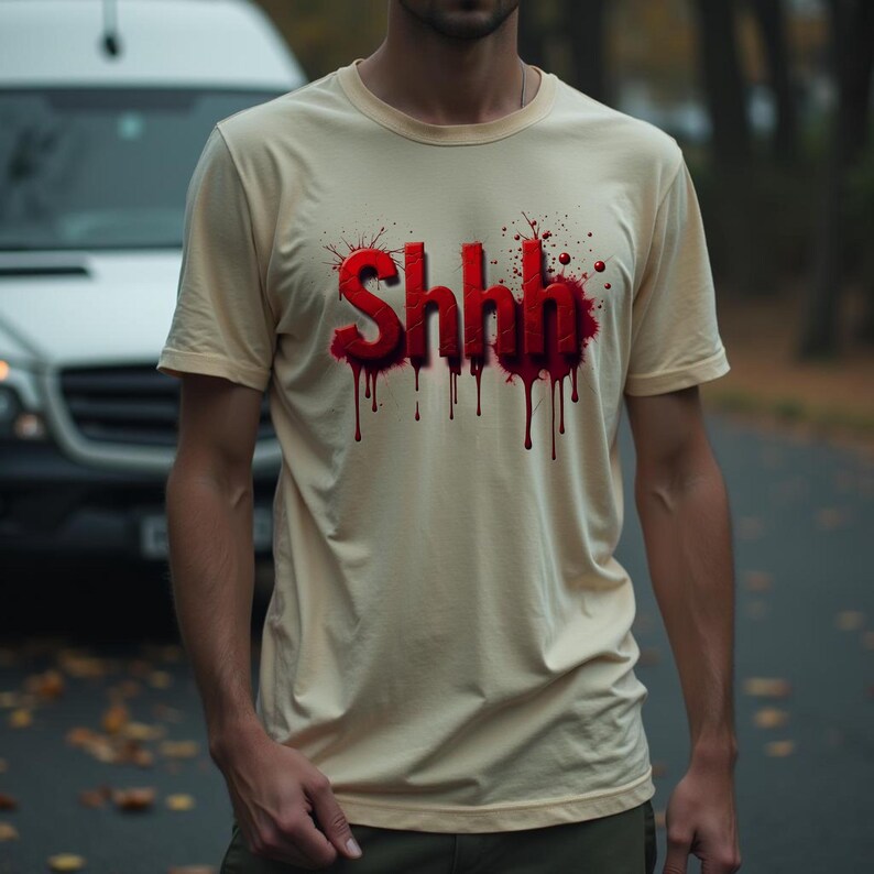 Pu&ograve; includere: T-shirt color crema con la scritta "Shhh" in rosso, con un effetto gocciolante simile a sangue. La maglietta a maniche corte ha uno scollo tondo. La persona indossa la maglietta all'aperto.
