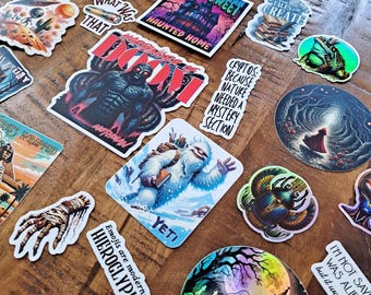 May include: A collection of colourful stickers featuring various designs, including a monster, a yeti, a haunted house, a frog, a creature from the black lagoon, and a ghost. The stickers are on a wooden surface.