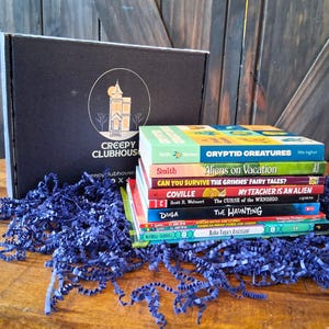 May include: A black cardboard box with the white text Creepy Clubhouse and a house illustration, accompanied by a stack of children's books including titles like Cryptid Creatures, Aliens on Vacation, and The Haunting, all resting on blue paper shreds.