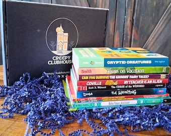 May include: A black cardboard box featuring the white text Creepy Clubhouse and a house illustration, accompanied by a stack of children's books including titles like Cryptid Creatures, Aliens on Vacation, and The Haunting, all resting on blue paper shreds.