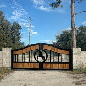 May include: A double-swing driveway gate featuring a black metal frame with horizontal wood-grain panels and a central circular medallion containing a silhouette of a rearing horse, set between two light-colored stone masonry pillars.