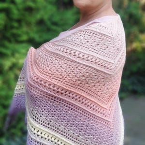 May include: A crocheted shawl in a gradient of colors, including lavender, peach, and cream. The shawl is draped over the shoulders, showcasing the intricate stitch pattern and color transitions. The background is a blurred green.