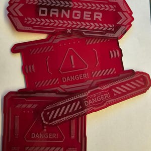 Könnte beinhalten: Drei rote Warnschilder aus Kunststoff mit dem Wort "DANGER" in weißen Buchstaben. Die Schilder haben ein futuristisches, geometrisches Design mit diagonalen Linien und Pfeilen.