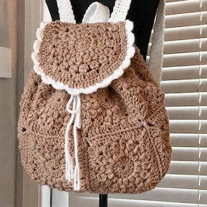 Peut inclure: Sac à dos en crochet marron avec un motif floral. Le rabat et les bretelles sont blancs, créant un contraste. Le sac a une fermeture à cordon et est présenté sur un support noir. Un accessoire unique et fait à la main.