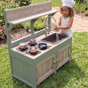 Pode incluir: Uma cozinha de brinquedo de madeira com estrutura verde sálvia, pia marrom com torneira prateada e bancada com três queimadores pretos. Inclui duas panelas cor de cobre, uma frigideira preta e três utensílios de cozinha de metal pendurados.
