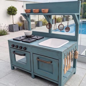 Könnte beinhalten: Eine blaue Spielküche aus Holz mit einem weißen Waschbecken, vier schwarzen Herdplatten und vier Drehknöpfen. Das Gerät verfügt über ein oberes Regal mit drei kleinen Schalen, hängenden Metall-Küchenutensilien und einen unteren Schrank.