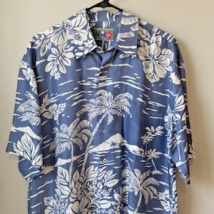 May include: Blue button-down shirt with a white tropical floral and palm tree print. The short-sleeved shirt is hanging on a black hanger. The shirt has a classic collar and a button-up front.