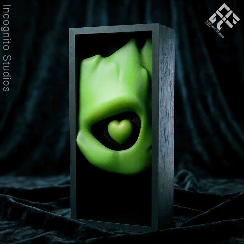 May include: A green sculpture of a face with a heart-shaped eye, set against a black background within a dark wooden frame. The sculpture has a smooth texture and is lit to highlight its form. The words "Incognito Studios" are printed on the left side of the frame.