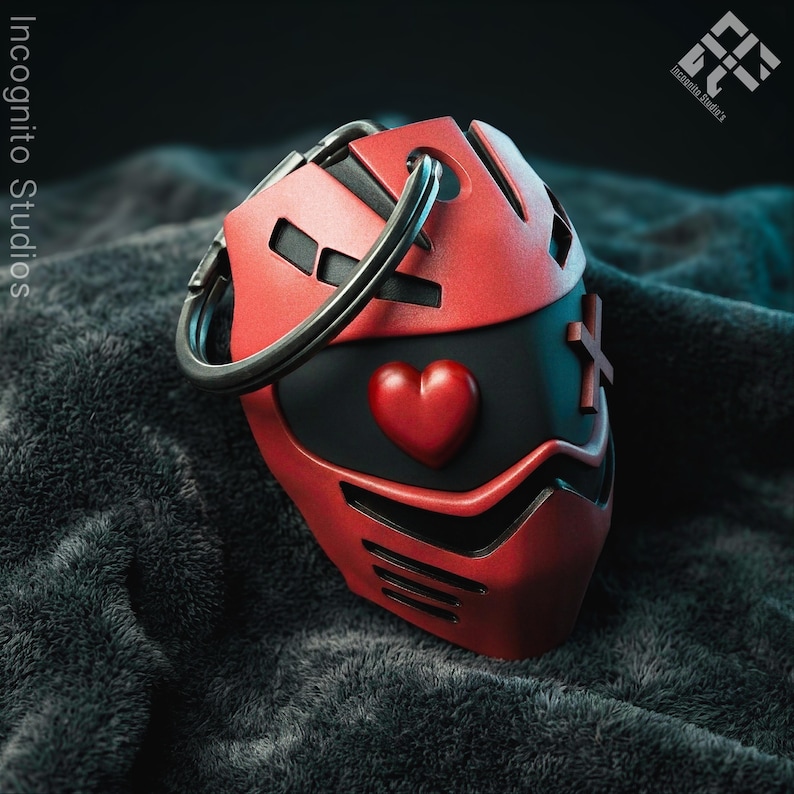 May include: A red and black keychain featuring a stylized mask design. The mask has a heart emblem and cross details. A silver key ring is attached to the top. The Incognito Studios logo is visible in the background.