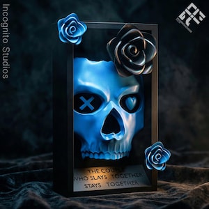 May include: A blue skull sculpture with an X and heart-shaped eye details, framed in black. Decorative blue and black roses adorn the frame. The text "THE COUPLE WHO SLAYS TOGETHER STAYS TOGETHER" is at the base.