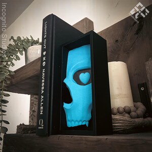May include: A black book titled "LIGHTS OUT" by Navessa Allen stands next to a framed, light blue skull with a heart-shaped eye. The skull is displayed in a black frame. A white candle and decorative beads are also visible on a wooden shelf.