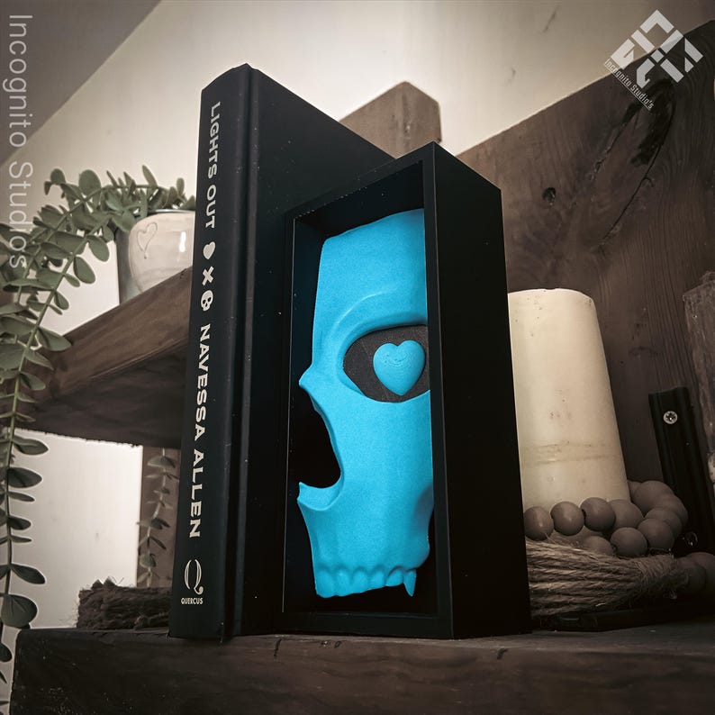 May include: A black book titled "LIGHTS OUT" by Navessa Allen stands next to a framed, light blue skull with a heart-shaped eye. The skull is displayed in a black frame. A white candle and decorative beads are also visible on a wooden shelf.