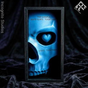 May include: A framed art print featuring a blue skull with a heart-shaped eye. The text reads "THE COUPLE WHO SLAYS TOGETHER STAYS TOGETHER." The artwork is set against a dark background, with the Incognito Studios logo in the top right corner.