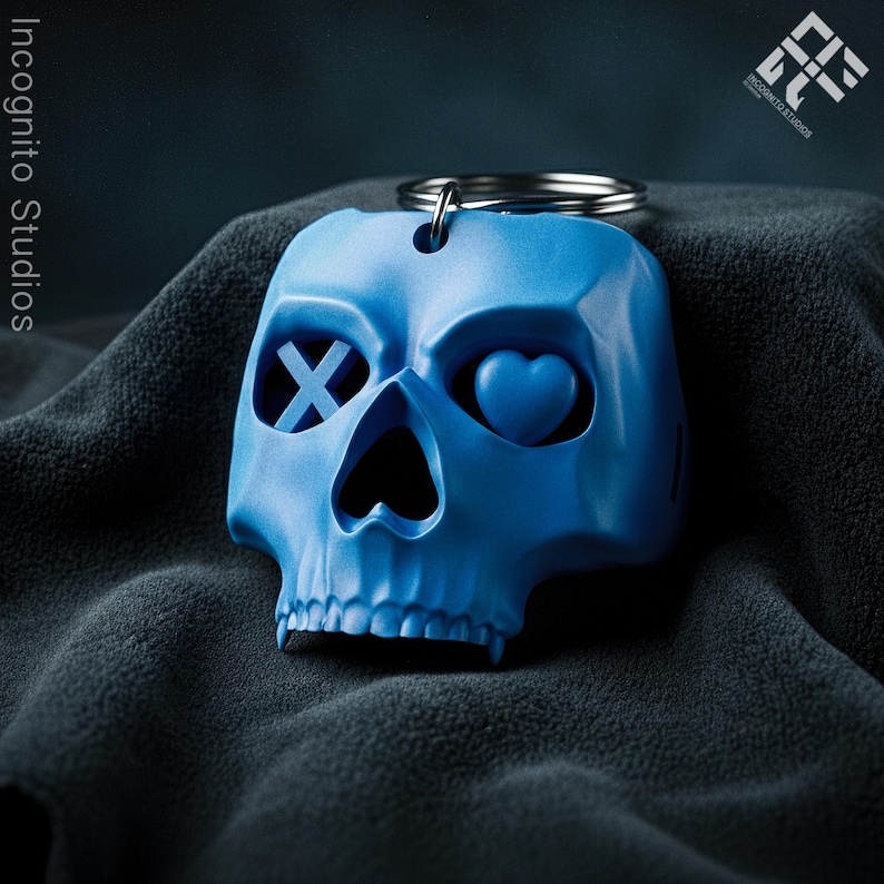May include: A blue skull-shaped keychain with a silver keyring. The skull has an X for one eye and a heart for the other. The keychain is made of a solid material and is a unique accessory.