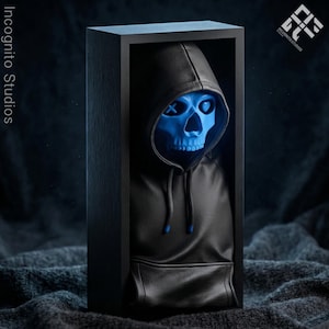 May include: A blue skull figure with an X and heart design on the eyes, wearing a black hoodie with a front pocket. The figure is displayed in a black wooden box. The Incognito Studios logo is visible.