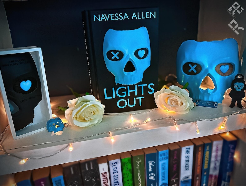 May include: A shelf displays a book titled "LIGHTS OUT" by Navessa Allen, featuring a blue skull design. A matching skull-shaped candle holder and other skull-themed decor are present, along with white roses and string lights. The shelf also holds a collection of books.