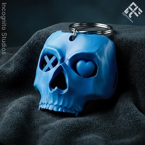 May include: A blue skull-shaped keychain with a silver keyring. One eye is an X, the other is a heart. The keychain is made of a solid material and is resting on a dark gray surface. The Incognito Studios logo is in the upper right corner.