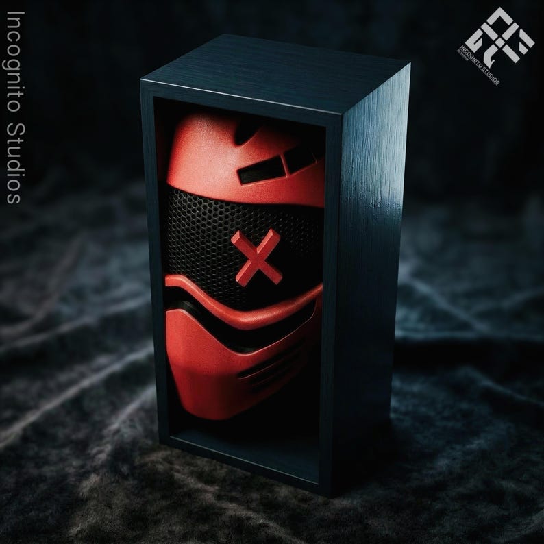 May include: A red and black mask with an X design is displayed inside a dark blue wooden box. The mask has a mesh front and is set against a dark, textured background. The box is rectangular and has a minimalist design.