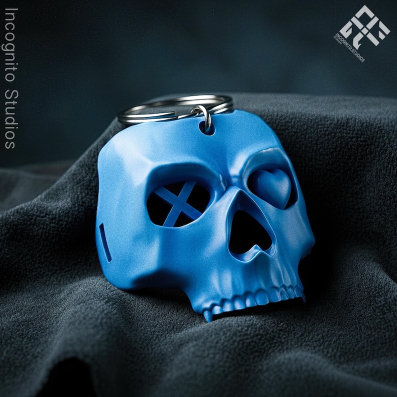 May include: A blue skull-shaped keychain with a silver keyring. The skull has heart-shaped eye sockets and an X-shaped design in one eye socket. The keychain is on a dark gray textured surface. The text "Incognito Studios" is visible.