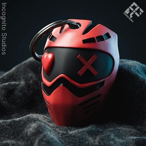 May include: A red and black keychain shaped like a stylized mask. The mask features a heart shape on one side and an X on the other. The keychain has a silver ring attached to the top. The words "Incognito Studios" are visible in the background.