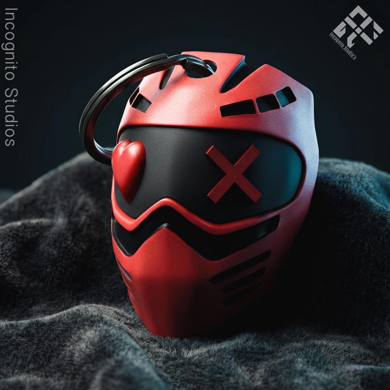 May include: A red and black keychain shaped like a stylized mask. The mask features a heart shape on one side and an X on the other. The keychain has a silver ring attached to the top. The words "Incognito Studios" are visible in the background.