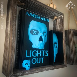 May include: A black book with the title "LIGHTS OUT" and author "NAVESSA ALLEN" in bright blue lettering. A blue skull graphic is on the cover. A small blue skull and a framed skull are also present. The items are displayed on a wooden shelf.