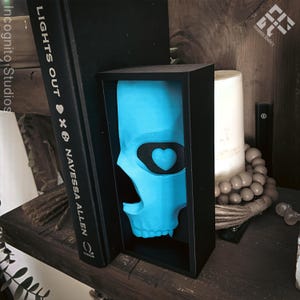 May include: A turquoise skull sculpture with a black heart-shaped eye patch is displayed in a black box. The sculpture is positioned between a black book with the title "LIGHTS OUT" and a white candle. The image is set on a wooden shelf.