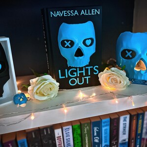 May include: A shelf display featuring a book titled "LIGHTS OUT" by Navessa Allen, with a blue skull design. The display includes a skull-shaped candle holder, a framed art piece, and a small figurine, all with a skull motif. White roses and string lights add to the aesthetic.
