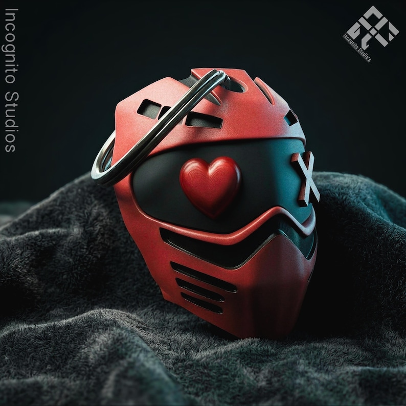 May include: A red and black mask with a heart and an X design. The mask has a silver ring on top and is set against a dark, textured background. The words "Incognito Studios" are visible.