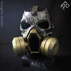 May include: A full-face mask featuring a weathered silver skull design with deep eye sockets and a central gold-coloured respirator unit. Two large, cylindrical gold filters with circular vents are attached to the sides. Text reads Incognito Studios.