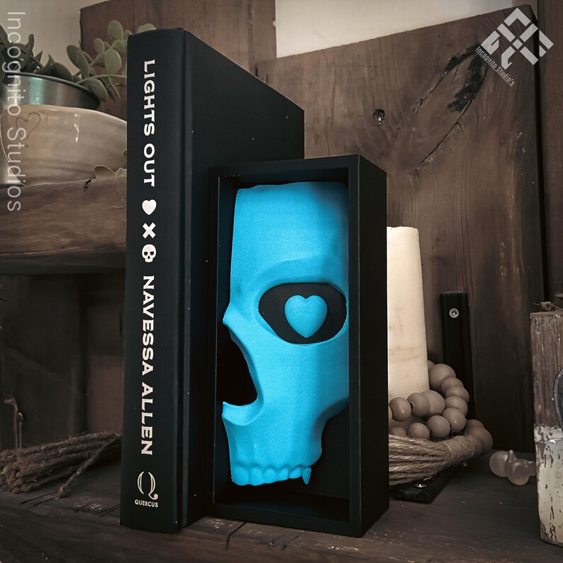 May include: A turquoise skull sculpture with a heart-shaped eye socket is displayed in a black shadow box. A black book with the title "LIGHTS OUT" and author "NAVESSA ALLEN" stands next to it. The scene is set on a wooden shelf.