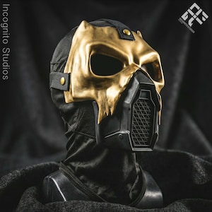 May include: A gold and black mask with a skull-like design. The mask features a metallic gold upper section and a black lower section with a geometric pattern. The mask is displayed on a black surface.