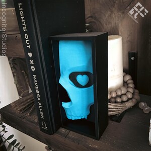 May include: A turquoise skull sculpture with a black heart-shaped eye patch is displayed in a black box. The sculpture is positioned between a black book with the title "LIGHTS OUT" and a white candle. The image is set on a wooden shelf.