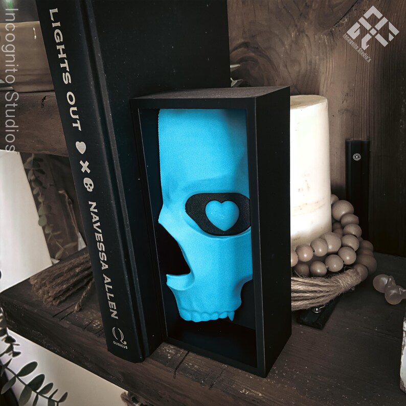 May include: A turquoise skull sculpture with a black heart-shaped eye patch is displayed in a black box. The sculpture is positioned between a black book with the title "LIGHTS OUT" and a white candle. The image is set on a wooden shelf.