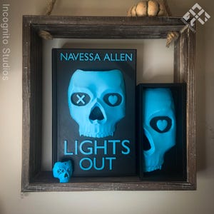 May include: A wooden shelf displays a book and two skull-shaped art pieces. The book cover is black with a blue skull design and the text "LIGHTS OUT" and "NAVESSA ALLEN". The skulls are light blue.
