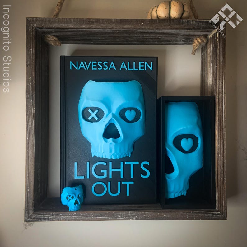 May include: A wooden shelf displays a book and two skull-shaped art pieces. The book cover is black with a blue skull design and the text "LIGHTS OUT" and "NAVESSA ALLEN". The skulls are light blue.