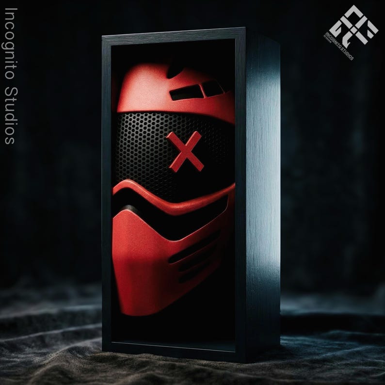 May include: A red and black decorative piece featuring a stylized mask design with an 'X' symbol. The mask is set against a black mesh background within a black frame. The words "Incognito Studios" are visible.
