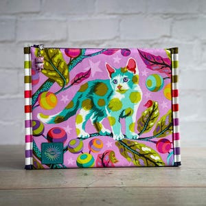 May include: A rectangular zippered pouch with a colorful cat design. The cat is teal with yellow spots, set against a pink background with stars, leaves, and colorful orbs. The sides have vertical stripes.