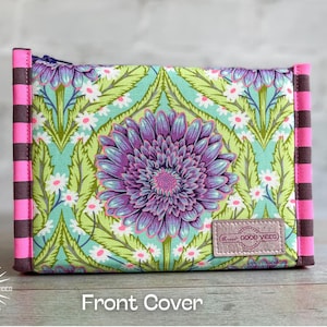 May include: A rectangular fabric pouch featuring a central purple flower on a light green background with small white flowers and green leaves. The sides have vertical dark brown and bright pink stripes. A small rectangular label reads GOOD VIBES.