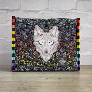 May include: A rectangular clutch featuring a detailed wolf illustration on a black background with colorful floral accents. The clutch has a rainbow-striped edge and a small, square label with text.