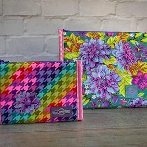 May include: Two zippered pouches. One has a colourful houndstooth pattern, the other a floral design with purple and blue flowers on a green background. Both have pink trim and a small label that says "GOOD VIBES".