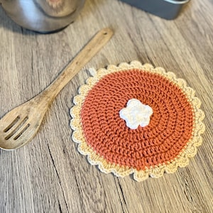 May include: A handmade, crocheted trivet in the shape of a pie. The trivet features a rust-colored center with a white, flower-shaped accent and a cream-colored, scalloped edge. A wooden spoon rests nearby.