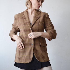 May include: A brown plaid blazer with a classic design. The blazer features a two-button closure, a notched lapel, and two front pockets. The plaid pattern includes shades of brown and red. The blazer is paired with a black skirt and off-white tights.