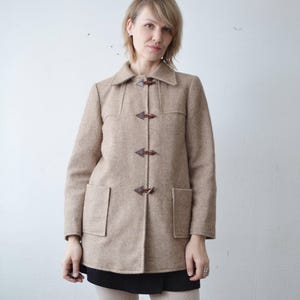 May include: A tan toggle coat with a collared neckline and two front pockets. The coat features three brown toggle closures and is made of a wool-like material. The coat is knee-length.