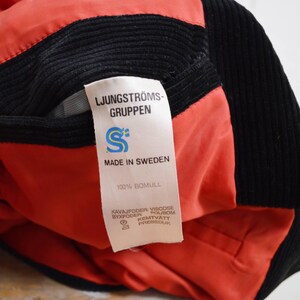 May include: Close-up of a black corduroy jacket with a red lining. A white tag displays the text "LJUNGSTR&Ouml;MS-GRUPPEN", "MADE IN SWEDEN", and "100% BOMULL". The jacket's interior is visible, showcasing the red fabric.