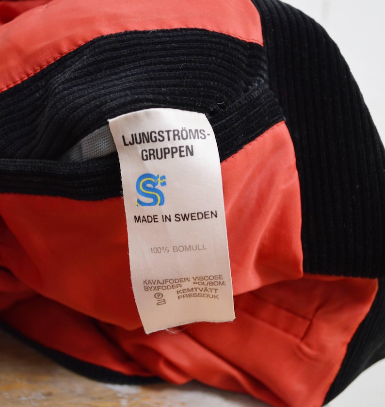 May include: Close-up of a black corduroy jacket with a red lining. A white tag displays the text "LJUNGSTR&Ouml;MS-GRUPPEN", "MADE IN SWEDEN", and "100% BOMULL". The jacket's interior is visible, showcasing the red fabric.