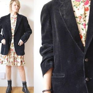 May include: A black corduroy blazer with a notched lapel and two-button closure. The blazer is worn over a floral print dress. The sleeves are long and the blazer has a classic, tailored fit. The outfit is completed with black boots.