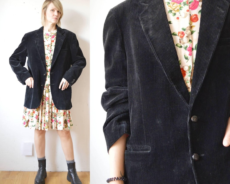 May include: A black corduroy blazer with a notched lapel and two-button closure. The blazer is worn over a floral print dress. The sleeves are long and the blazer has a classic, tailored fit. The outfit is completed with black boots.