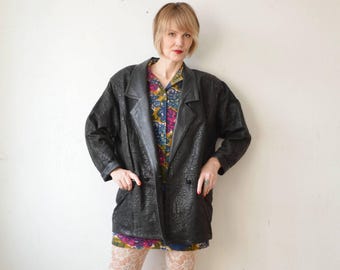 May include: A black leather jacket with a textured pattern. The jacket has a double-breasted closure and a collar. The jacket is worn over a floral print shirt.